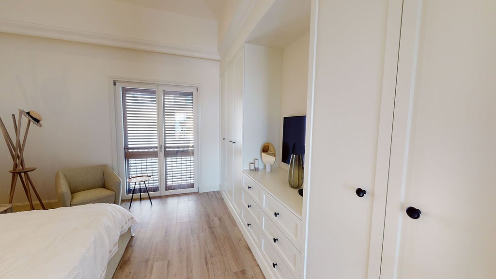 Bright bedroom in an apartment, Luštica, Montenegro: built-in wardrobe, chest of drawers, armchair, and balcony access with blinds.
