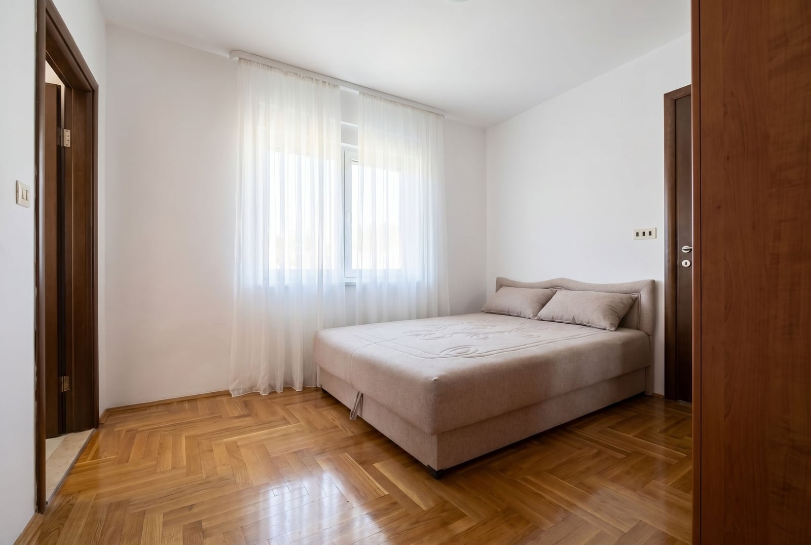 Bright bedroom with a double bed and parquet flooring in a house, Baošići, Montenegro. Interior with white walls and a window.