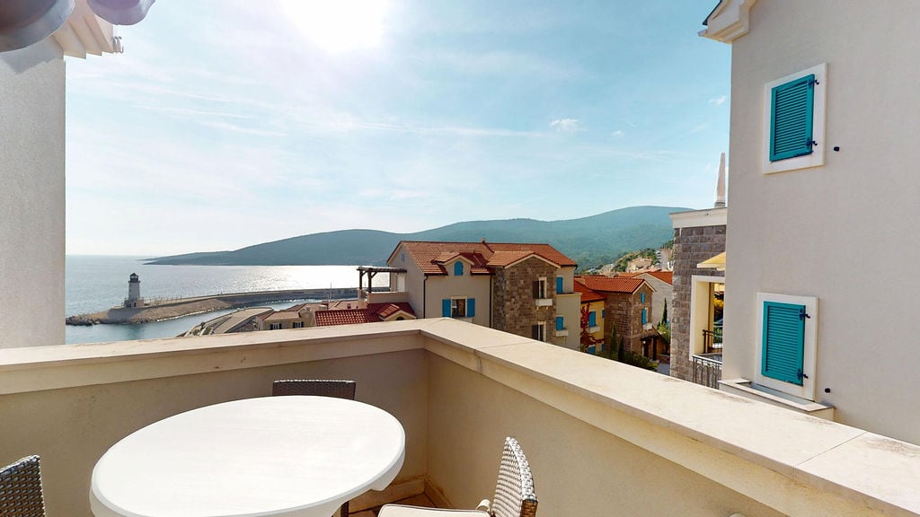 View from the balcony overlooking the sea, lighthouse, and mountains at the Luštica Bay residential complex, Krašići, Montenegro. A table and chairs are on the terrace.