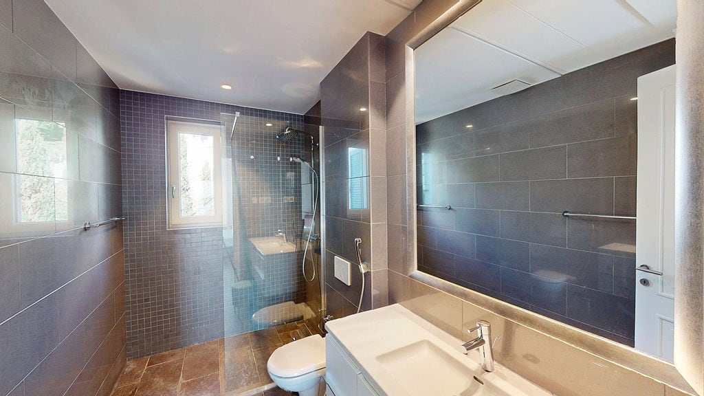 Modern bathroom with a shower cabin in an apartment, Luštica, Montenegro: gray tiles, large mirror, and plumbing fixtures.