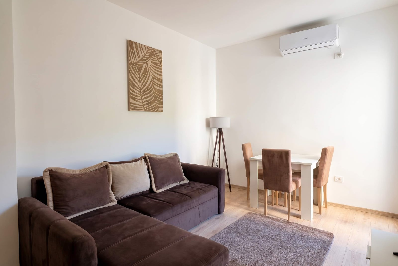 Bright living room in an apartment in Budva, Montenegro: brown corner sofa, dining area, and air conditioner.
