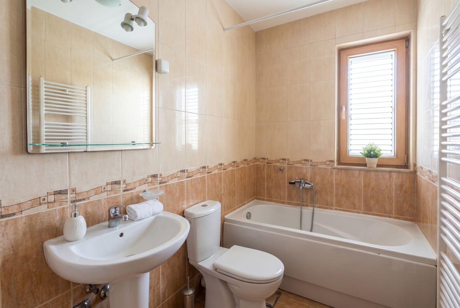 Bright bathroom with white fixtures and a window in the house, Baošići, Montenegro. Beige tiles, bathtub, and mirror.