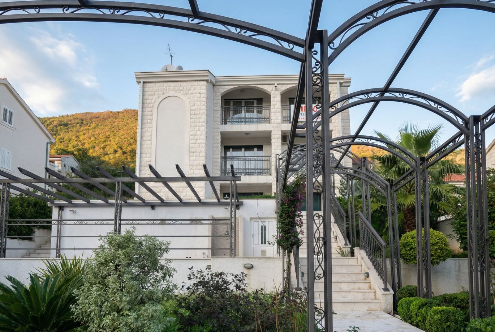 Three-story light stone house with terraces in Herceg Novi, Montenegro, with a wrought-iron pergola and a garden in the courtyard.