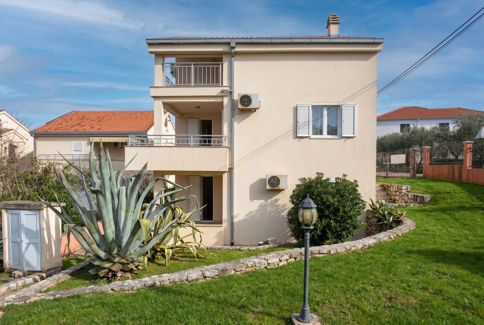 Three-story house in Baošići, Montenegro: facade with balconies, a well-kept green plot with agave and landscaping.