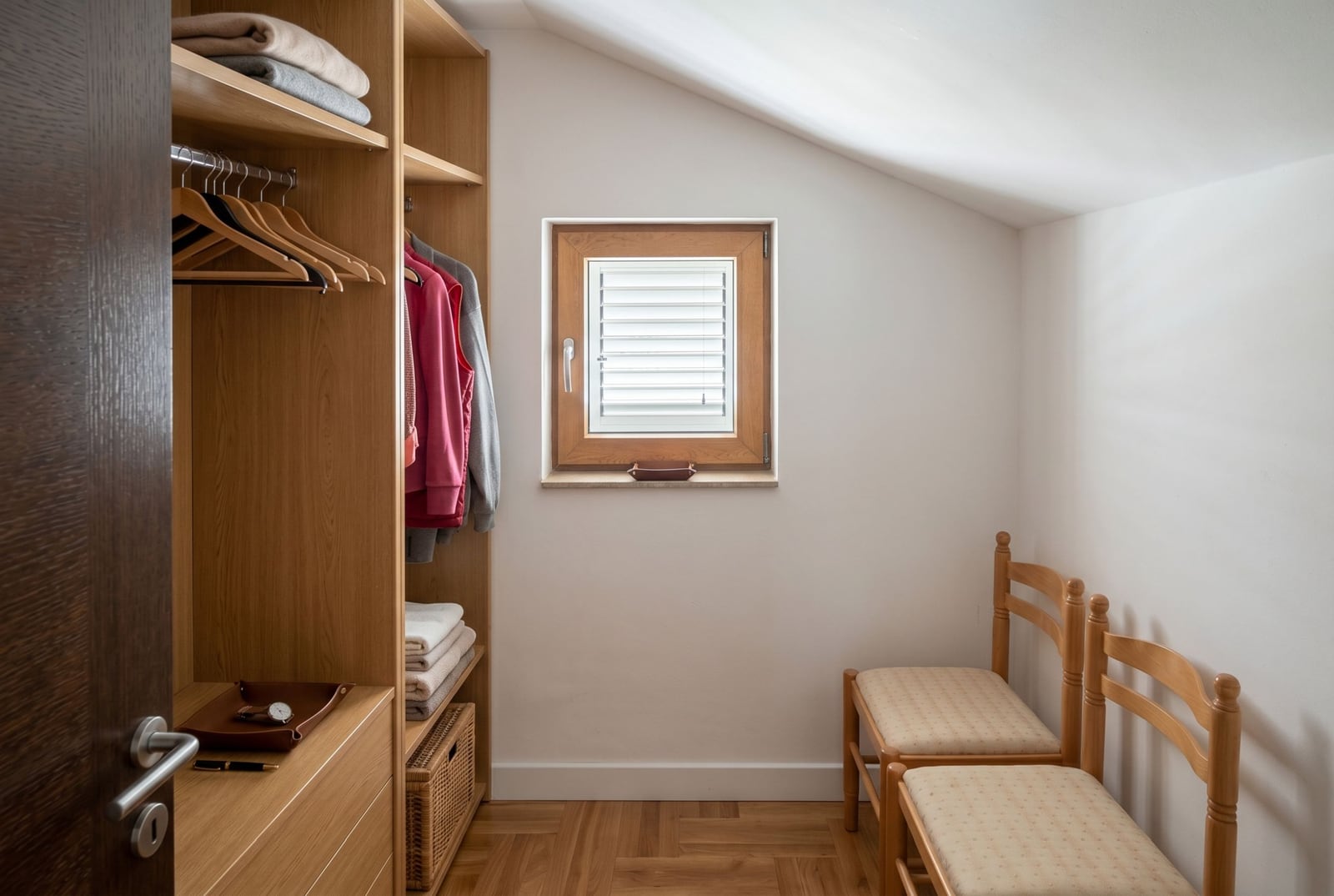 Walk-in closet with a wooden wardrobe and a window in a house, Baošići, Montenegro. Bright room with parquet flooring and two chairs.