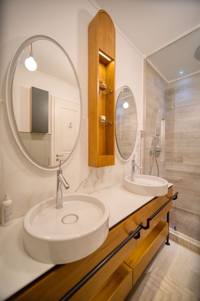Modern bathroom in an apartment, Luštica, Montenegro: two sinks, oval mirrors, and wood-like finish.