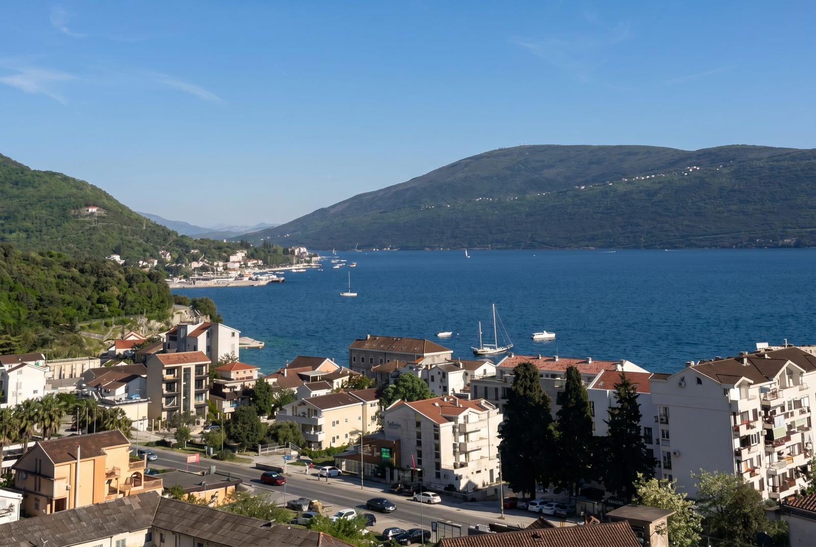 Panoramic view of Kotor Bay and the city of Herceg Novi, Montenegro: residential buildings, a coastal road, and yachts at sea.