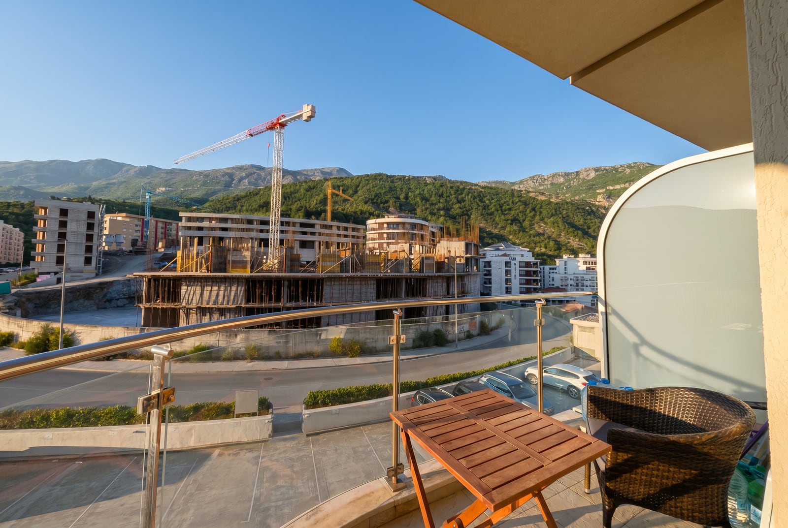 View from the balcony of an apartment in Bečići, Montenegro: wicker furniture, glass railing, and a panorama of mountains and construction.