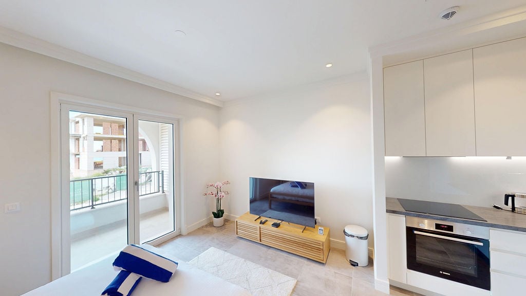 Modern living room with kitchen in Lustica Bay apartments, Montenegro: bright interior, TV area, and balcony access.