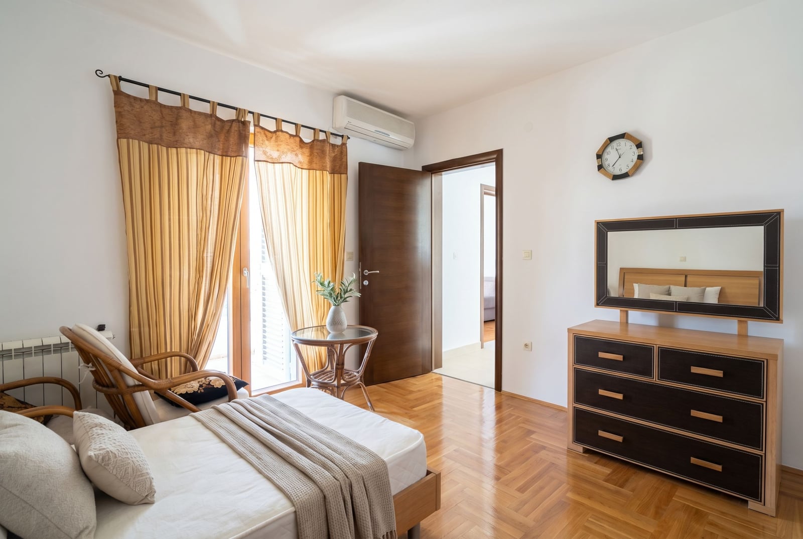 Bright bedroom in a house in Baošići, Montenegro: bed, rattan chair, dresser with mirror, and parquet floor.