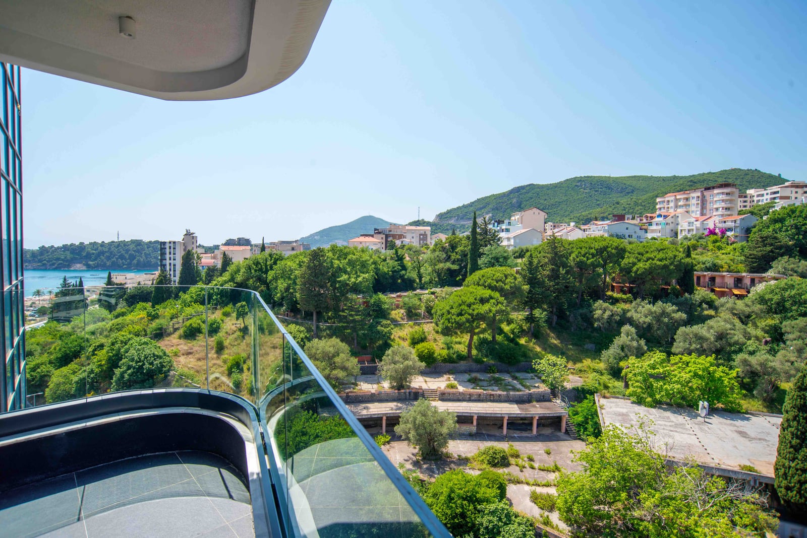 View from the balcony of a modern apartment in Bečići, Montenegro: a panorama of the sea, green hills, and urban development.