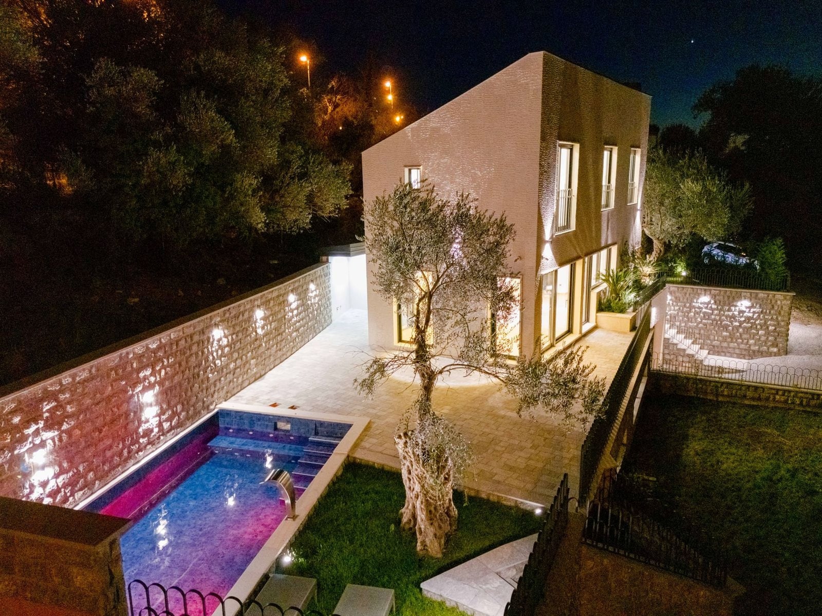 Villa moderne à Kamenovo, Monténégro : vue nocturne de la maison avec piscine, terrasse et olivier dans le jardin.