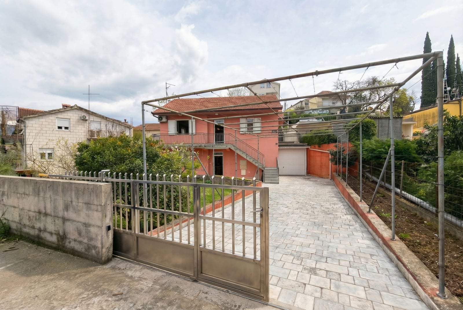 Two-story pink house in Herceg Novi, Montenegro, with a garage, paved courtyard, and a metal pergola for grapes.