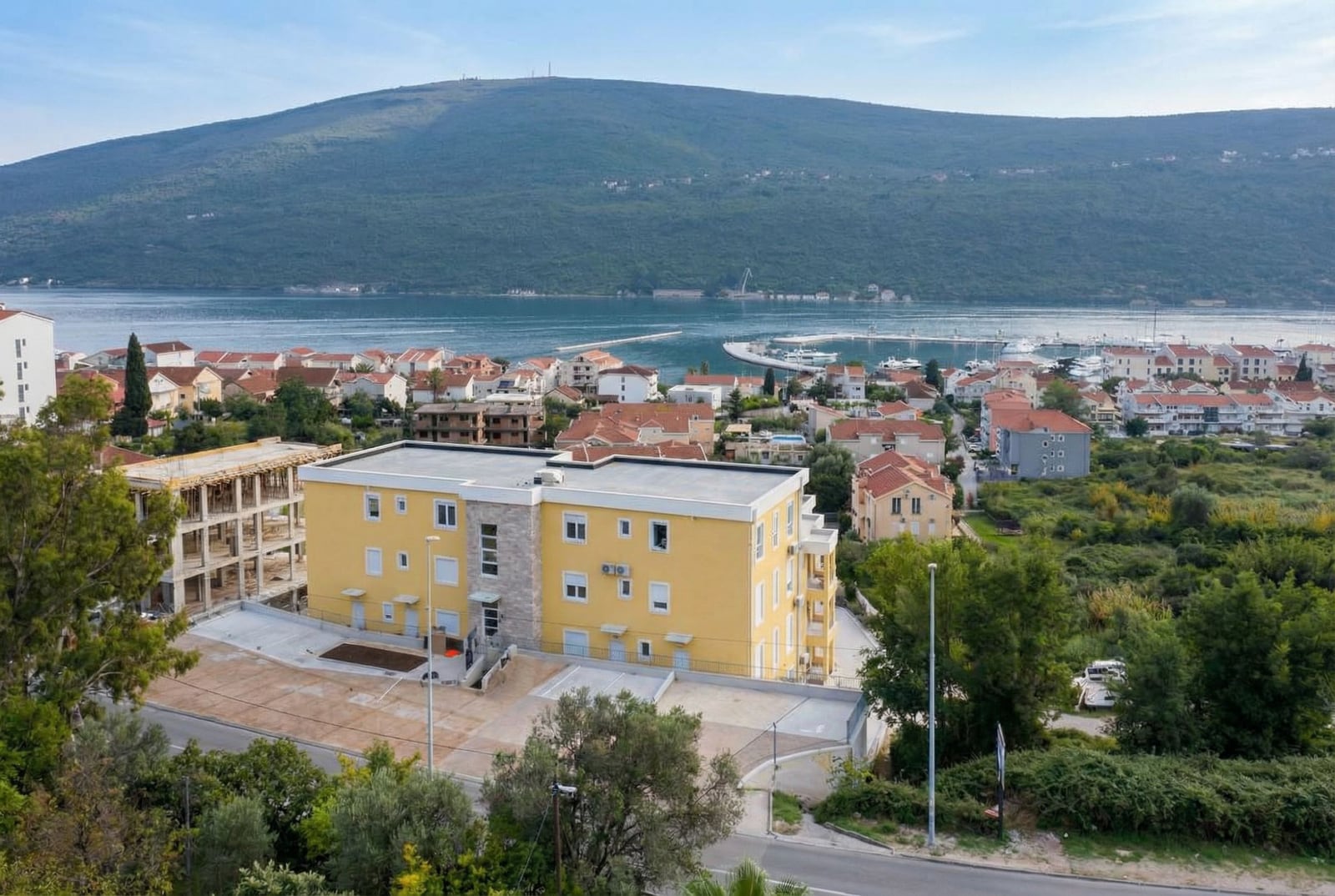 Aerial view of a yellow residential building in Herceg Novi, Montenegro, with panoramic views of the sea, mountains, and yacht marina.
